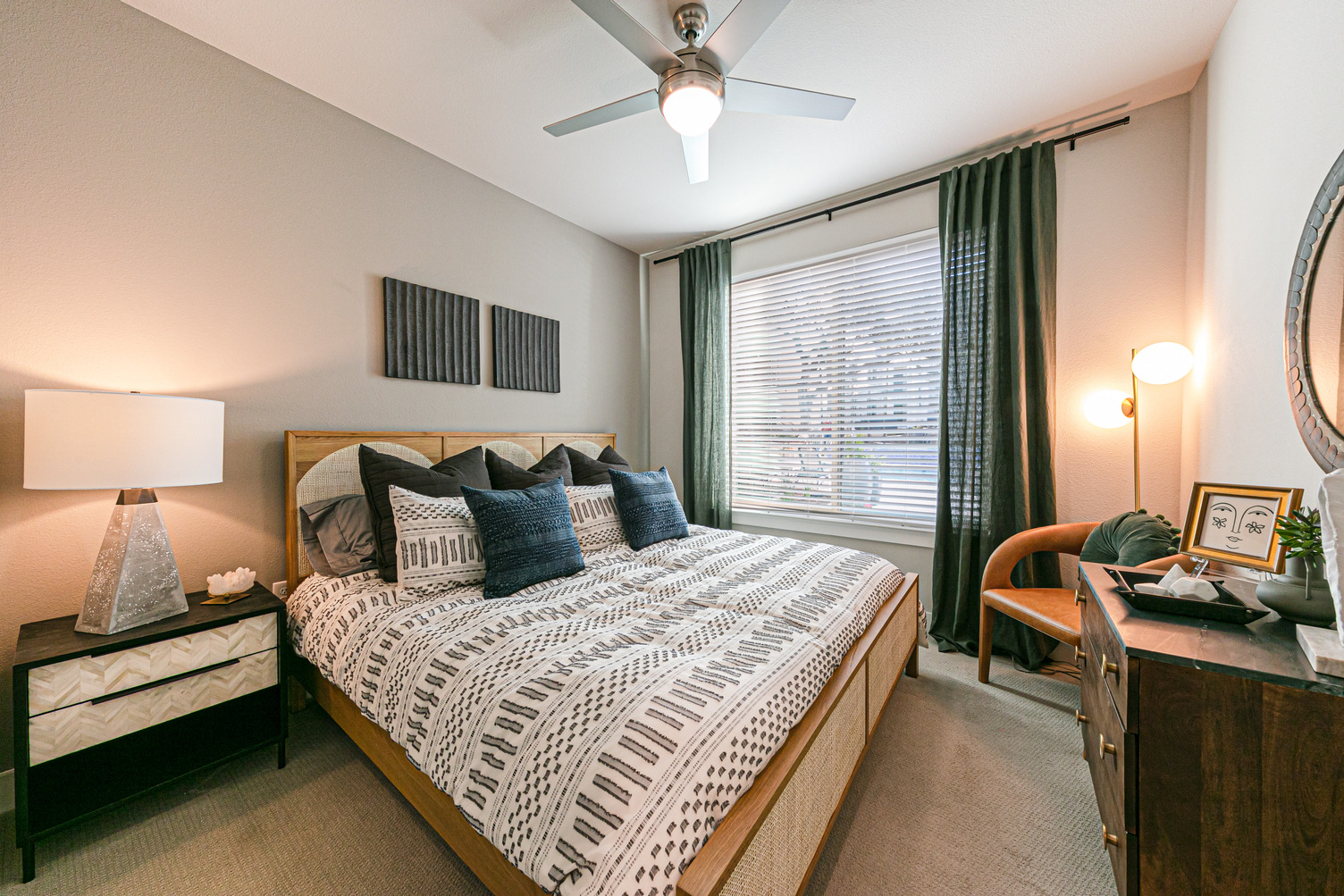 Model apartment bedroom with carpeting, ceiling fan, queen bed, and large window with blinds