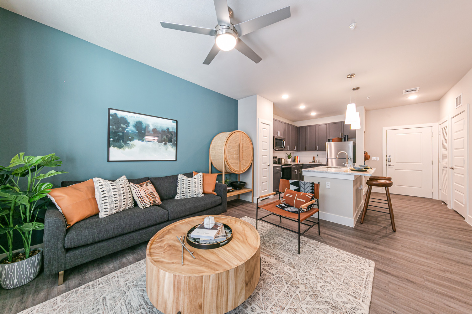 Model apartment interior viewed from living room with ceiling fan and open concept kitchen with island and wood plank style flooring throughout