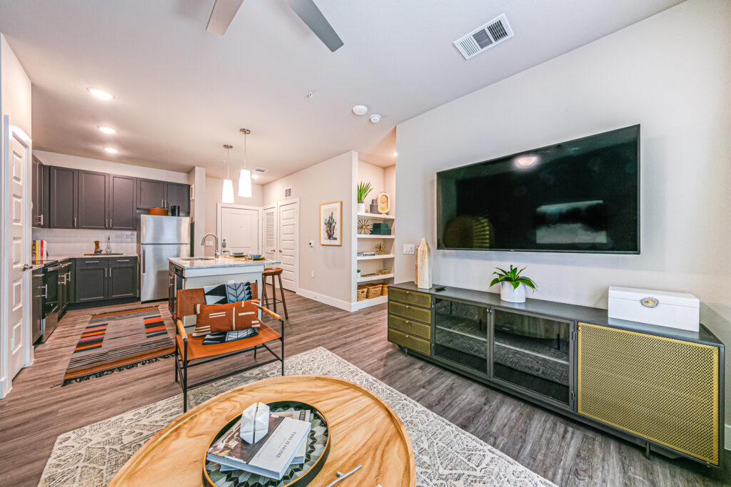 Model apartment with large TV console, built-in shelving, ceiling fan, recessed pendant lighting, and kitchen with island