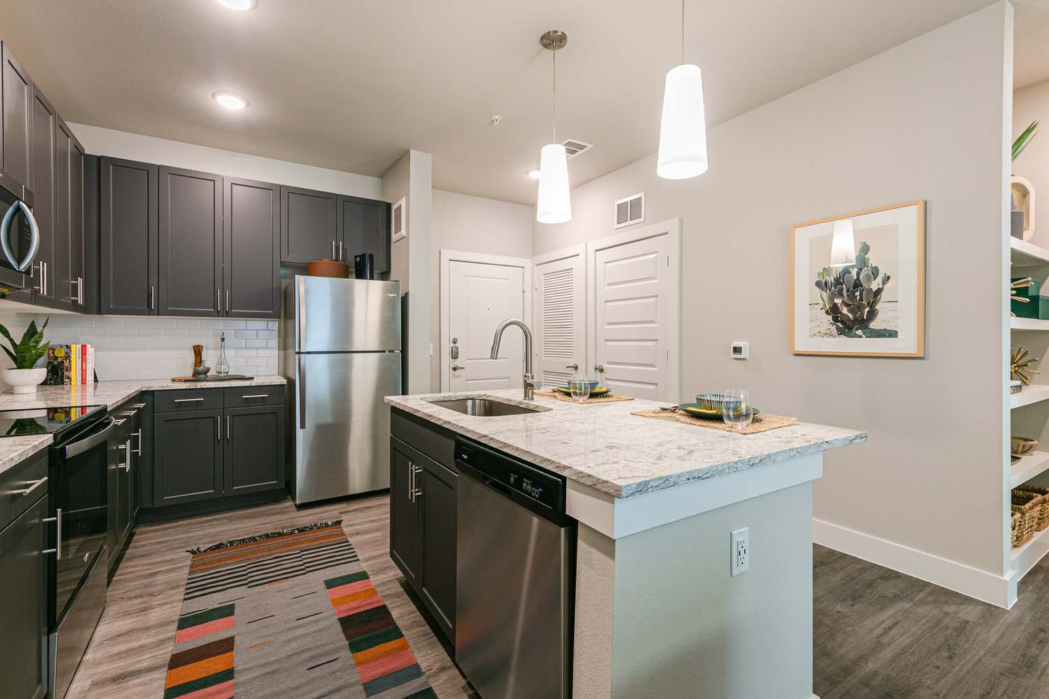 Model apartment kitchen with dark cabinets, quartz countertops, stainless steel appliances, and island with sink and dishwasher